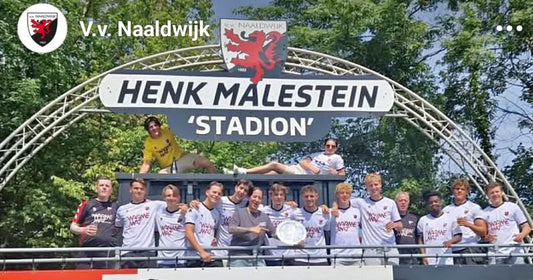 Een sfeervolle actiefoto van het team in hun De WarmeMat tenues op een zonnig Sportpark De Hoge Bomen, met de kampioensschaal prominent in beeld.